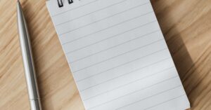 From above of small opened notepad and stylish silver pen placed on wooden table near laptop